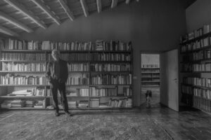 Blanco y negro. Foto de Jacobo Siruela. Está en su biblioteca de pie. Tras él, librerías colmadas de libros y una puerta por la que entra un perro