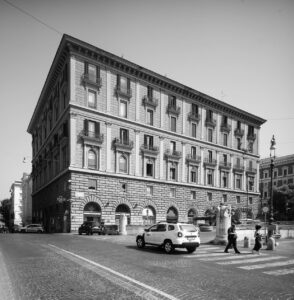 Foto urbana de Roma en blanco y negro. Un edifico en perspectiva, de arquitectura clásica. En primer término, un coche blanco aparcado y dos personas cruzando un paso de peatones.