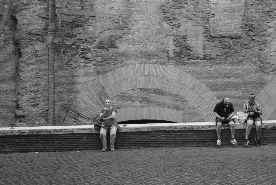 Blanco y negro. Foto de pared de ladrillos con arco romano en su base. En primer término tres turistas descansan sentados en un pequeño muro